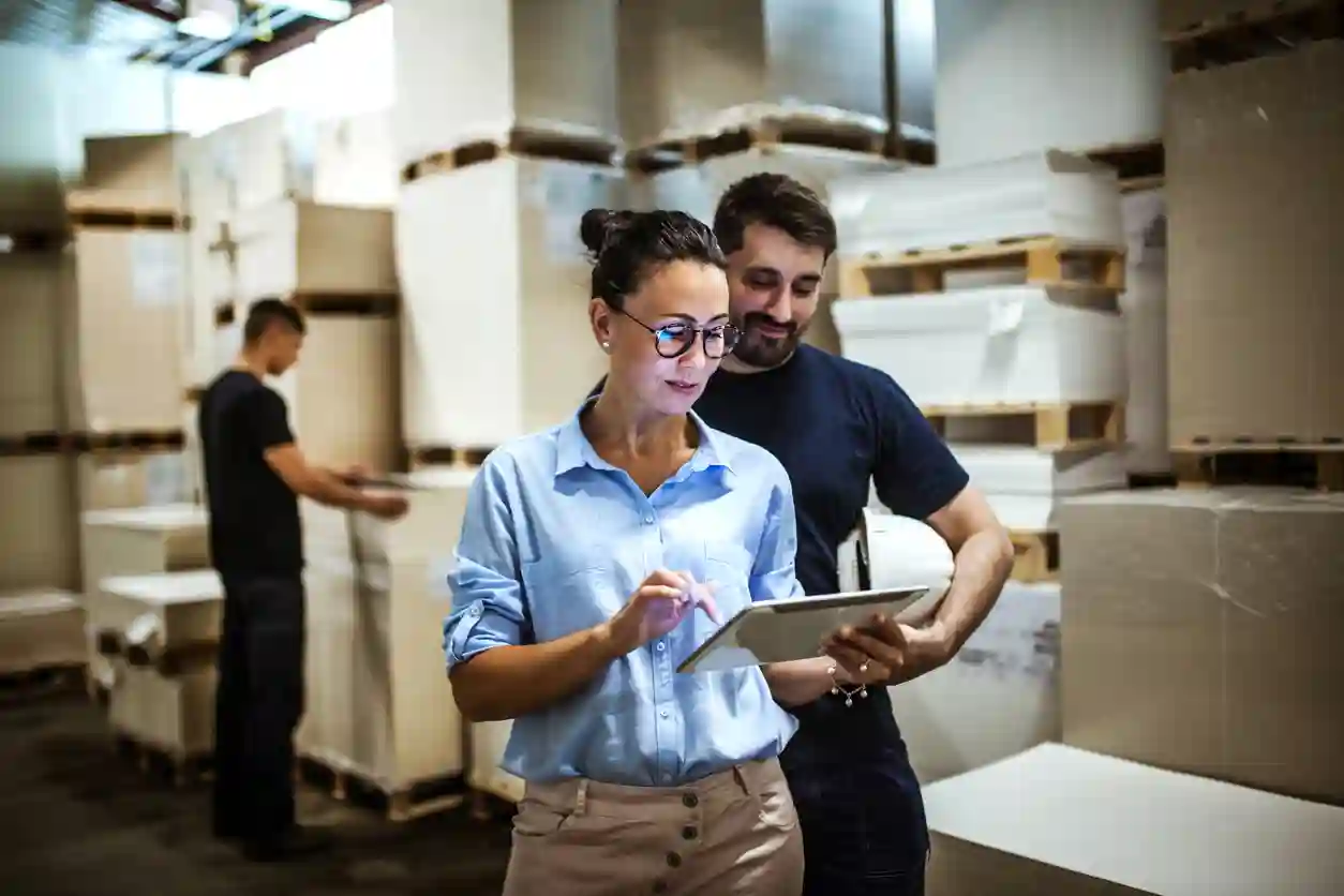 Two people look at a tablet in a warehouse wiht many boxes 